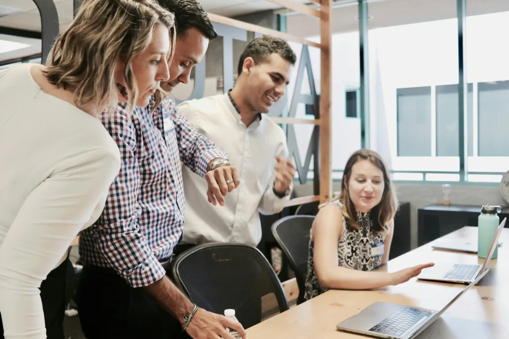 Four office colleagues lean over a wooden table while reviewing work together on a laptop in a modern workplace setting. (124 characters)