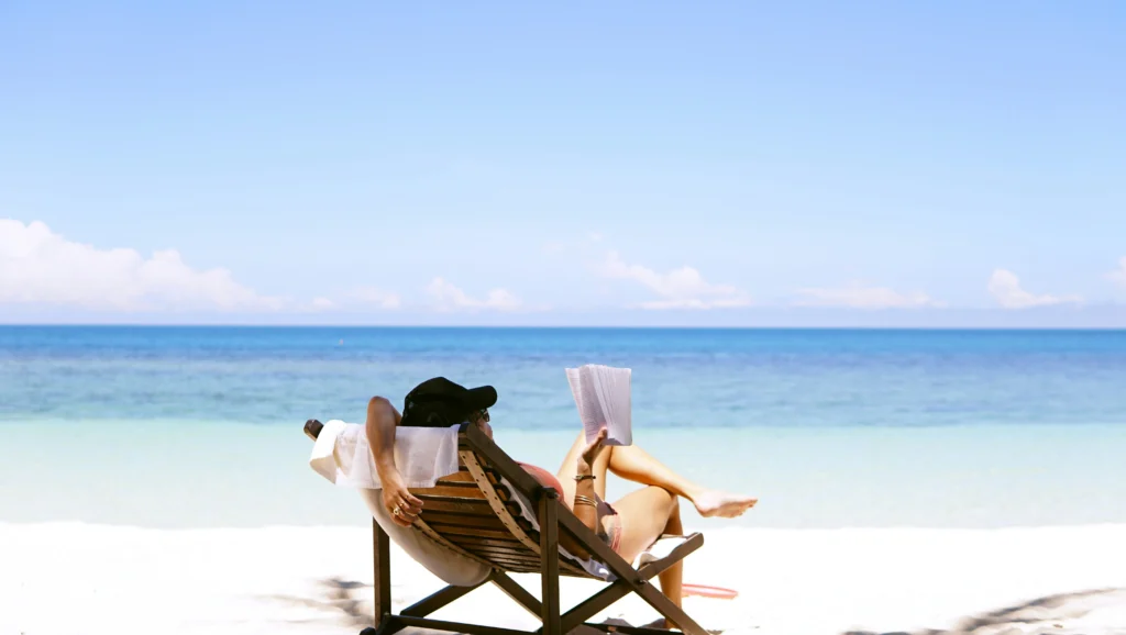 Two people relax in beach chairs beneath a bright blue sky, reading by turquoise ocean waves on a peaceful white-sand beach.