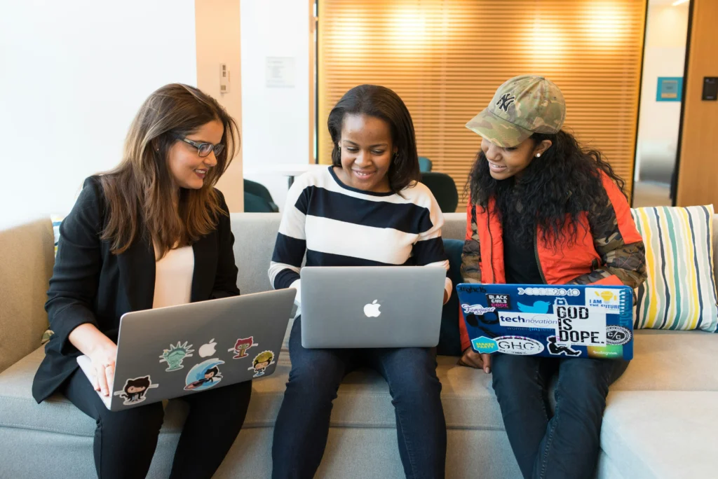 Three women sitting together on an office couch, smiling while working collaboratively on laptops adorned with sticker decorations.