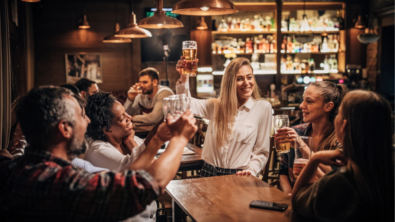 A group of friends raise their beer glasses in a cheer while enjoying drinks together inside a dimly lit bar.