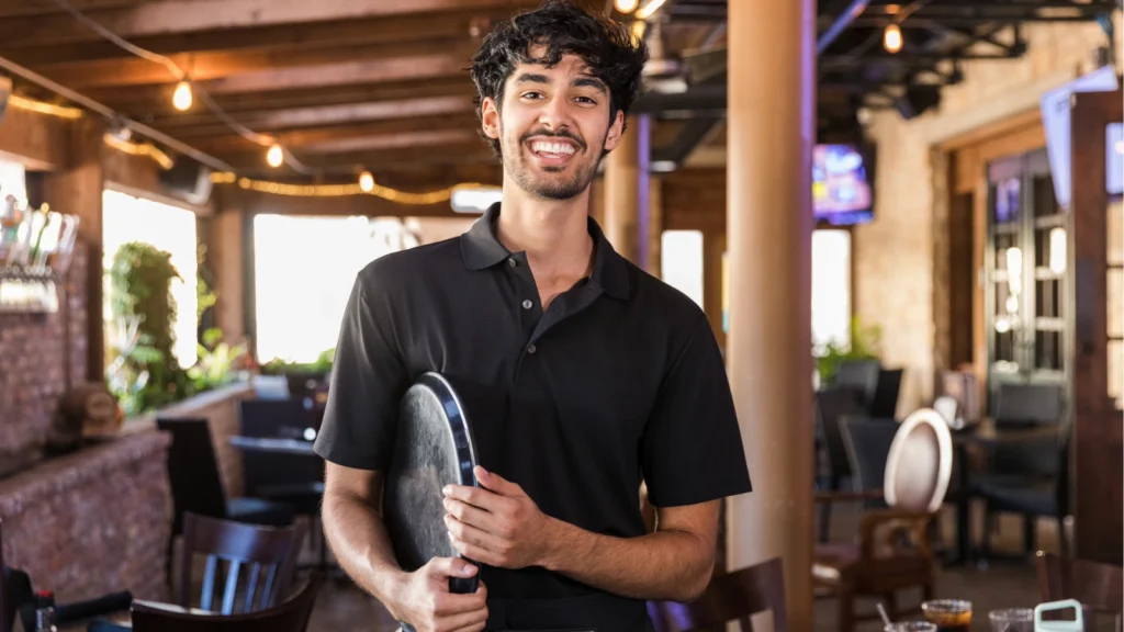 A smiling waiter wearing a black polo shirt stands inside a restaurant while holding a serving tray.