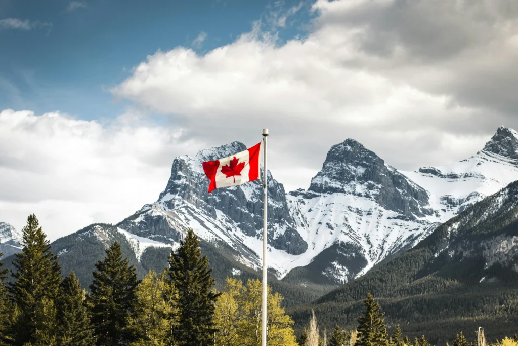 The Canadian flag flies proudly on a flagpole set against a breathtaking landscape of pine trees and snow-capped mountain peaks.