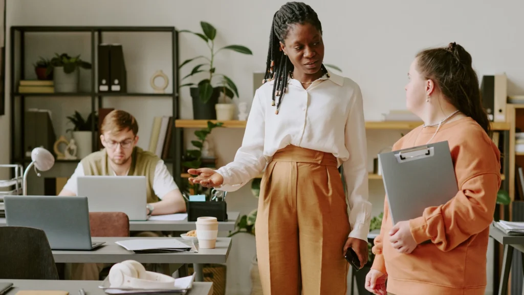 Diverse office team collaborates: a Black woman gestures while speaking with a colleague holding a tablet; a man works at a laptop.