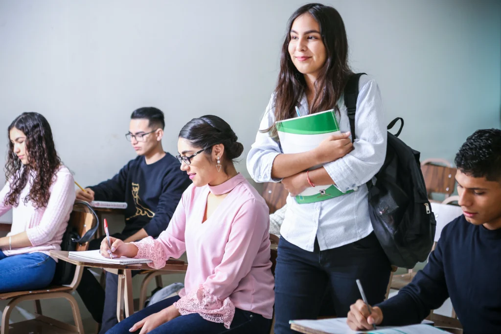 A bright classroom scene featuring several students sitting at individual desks and writing in notebooks. In the center, a smiling young woman stands up, holding a green binder or notebook close to her chest.
