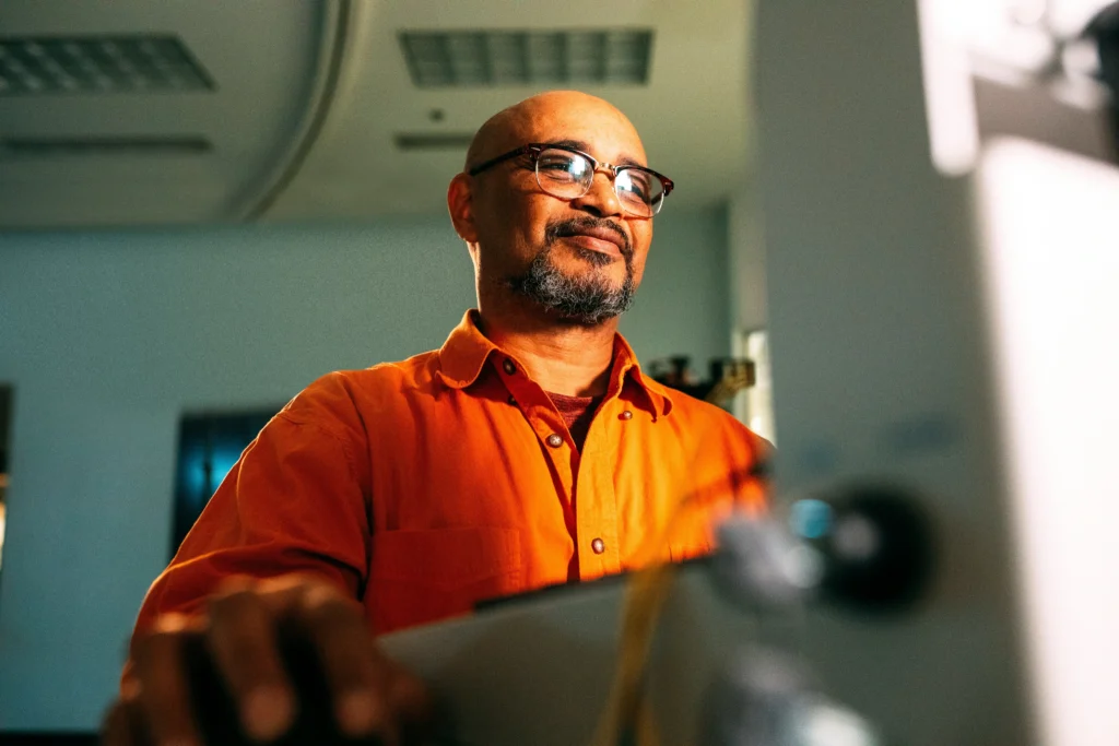 A low-angle shot of a smiling man with a goatee and glasses, wearing an orange shirt while focused on working on his desktop computer.