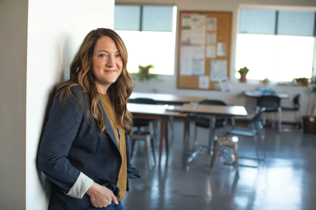 A confident female professional smiling warm-heartedly while leaning against a white pillar in a bright modern open-plan office space.