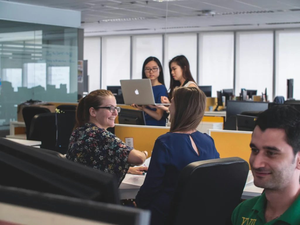 Diverse office employees talk warmly at desks in a open workplace setting while two women behind them review a laptop screen together.