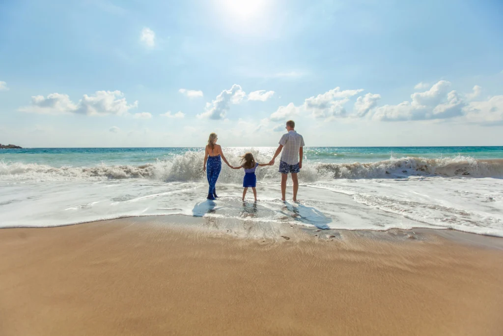 Family holding hands at the shoreline as waves roll in on a sunny beach, with turquoise ocean, blue sky, and a joyful vacation mood.