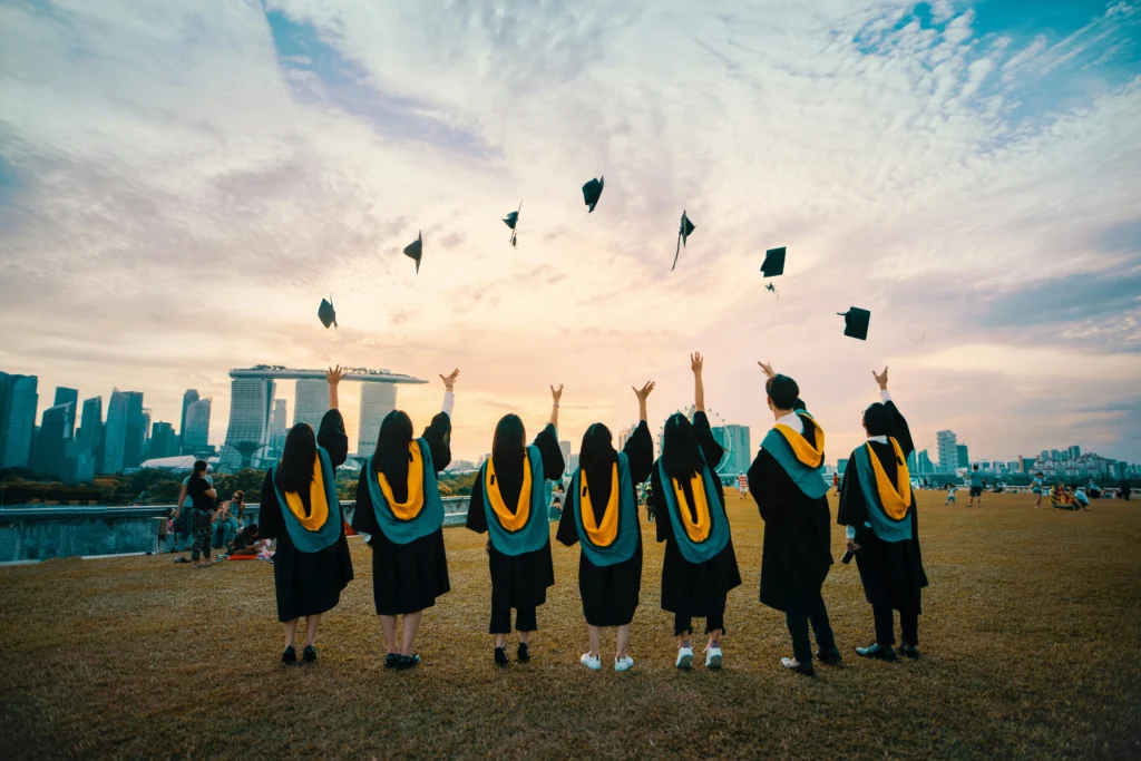 Seven university graduates seen from behind, wearing black graduation gowns with teal and yellow sashes, joyfully throwing their caps high into the air. They are standing on a grassy field with a colorful sunset and the Singapore city skyline, including Marina Bay Sands, in the background.