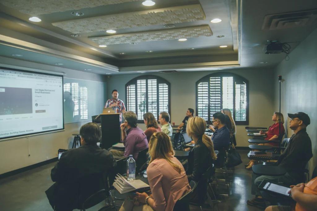 A presenter stands at a podium at the front of a room, speaking and displaying a slide presentation on a projector screen. A seated audience of about a dozen people is attentively listening and taking notes on chairs with attached desk arms.