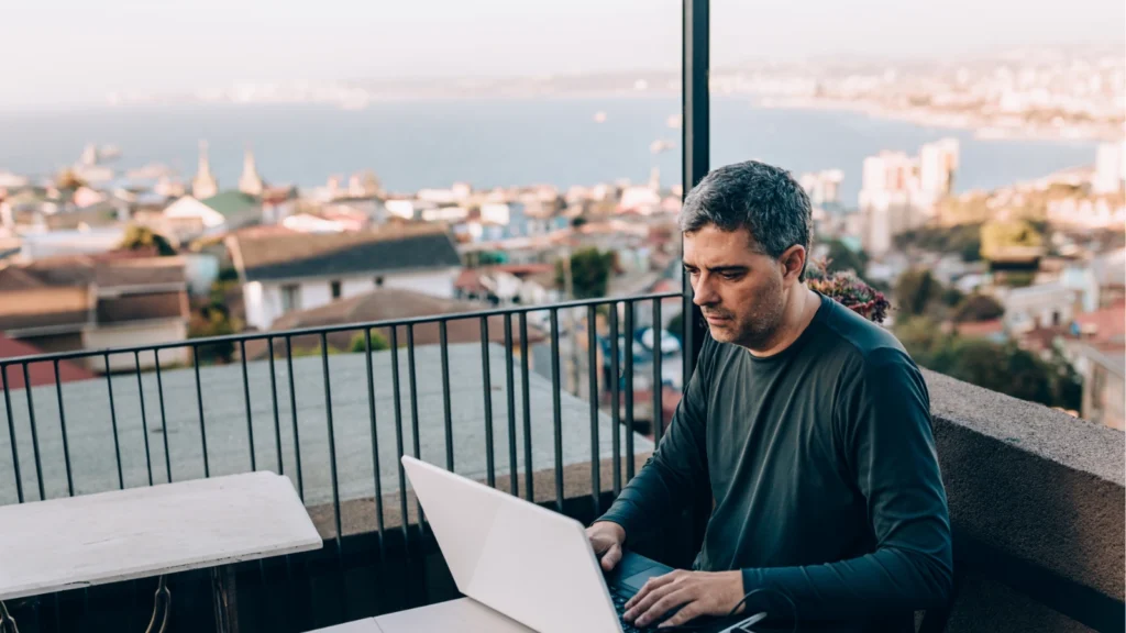 A middle-aged man works intently on his laptop computer while sitting on a outdoor balcony with a sweeping city and ocean view.