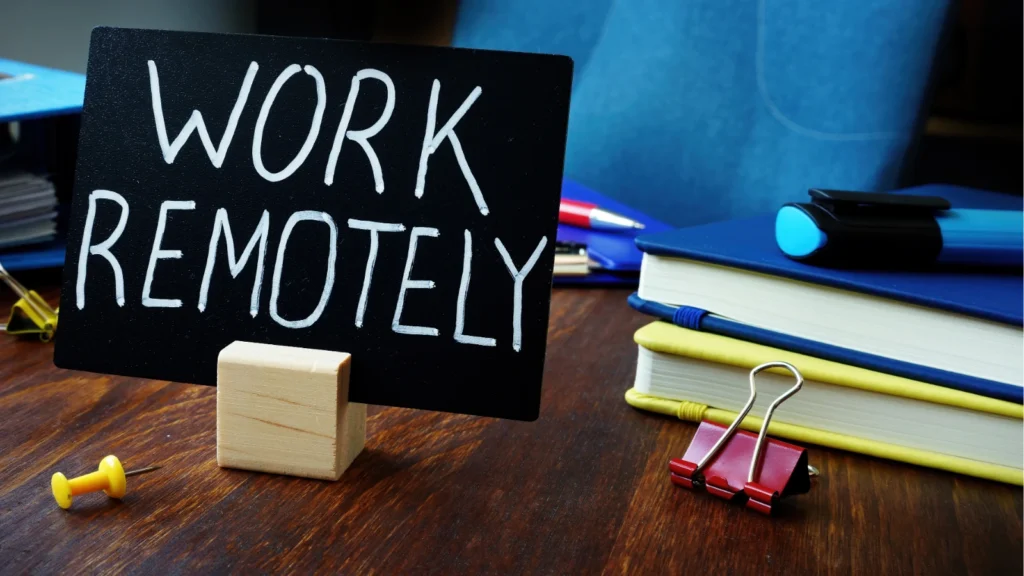 A small black sign reading "WORK REMOTELY" sits on a wooden desk alongside notebooks, a highlighter, and office supplies.