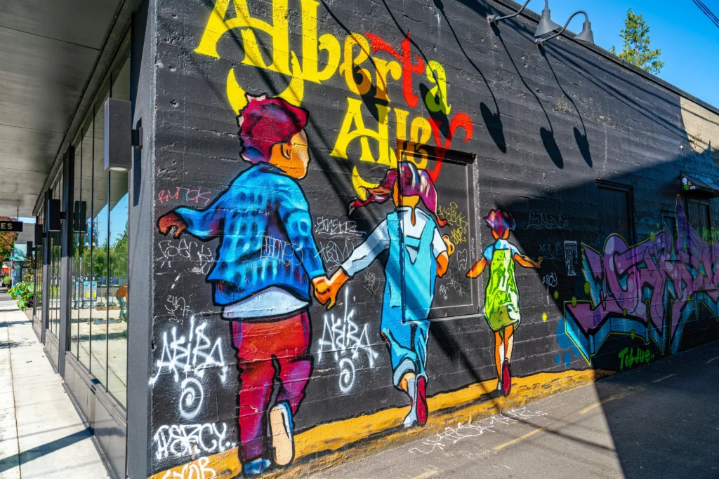 A colorful street art mural painted on a black building wall depicts children holding hands below bold lettering reading "Alberta Alley."