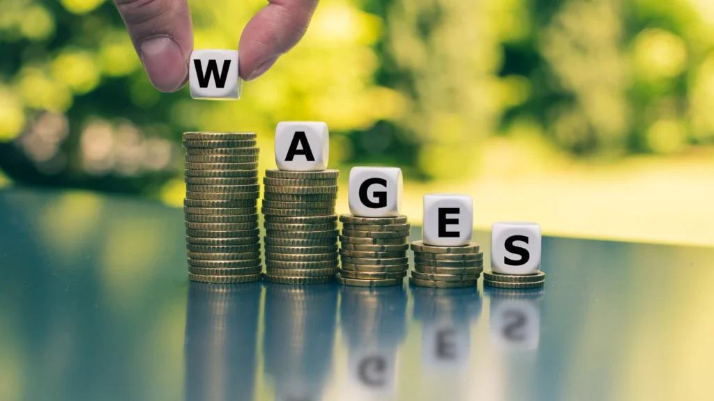 A hand places lettered letter blocks spelling "WAGES" onto declining stacks of gold coins set against a blurred green park background.
