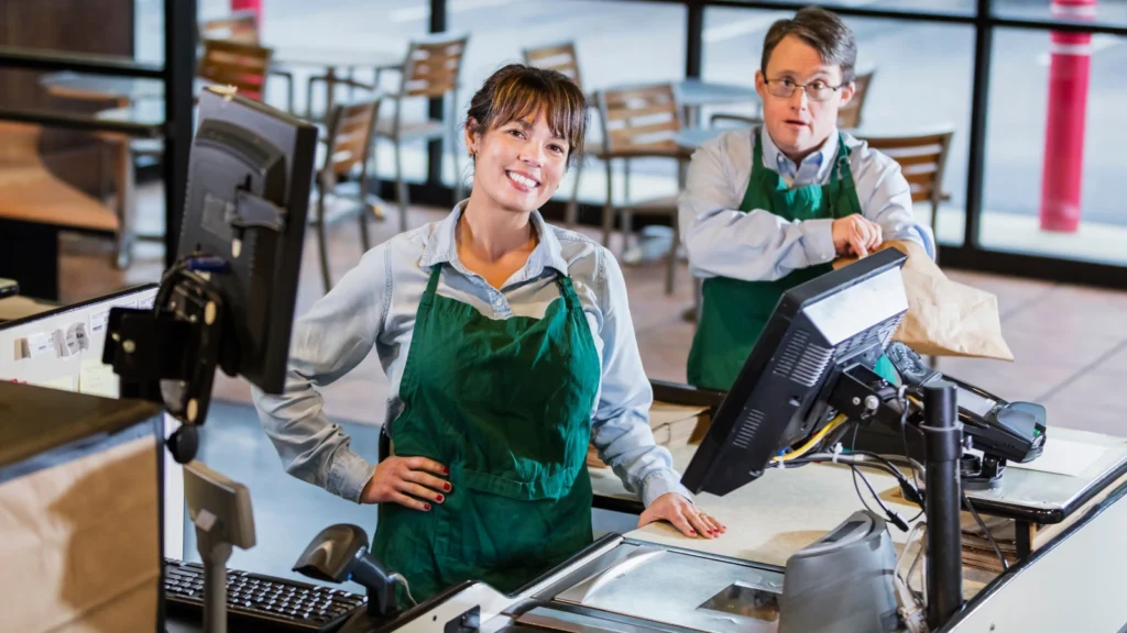 A female cashier in a green apron smiles warmly behind a grocery checkout counter while her male colleague packs groceries nearby.