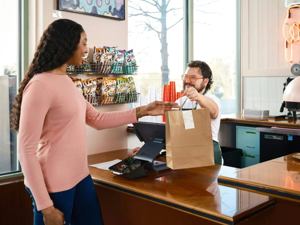 A smiling woman in a pink long-sleeved shirt receives a brown paper takeout bag from a cashier wearing glasses behind a wooden counter. The shop's interior includes a point-of-sale system on the counter, wall-mounted racks displaying bags of chips, and large windows showing bare trees outside.