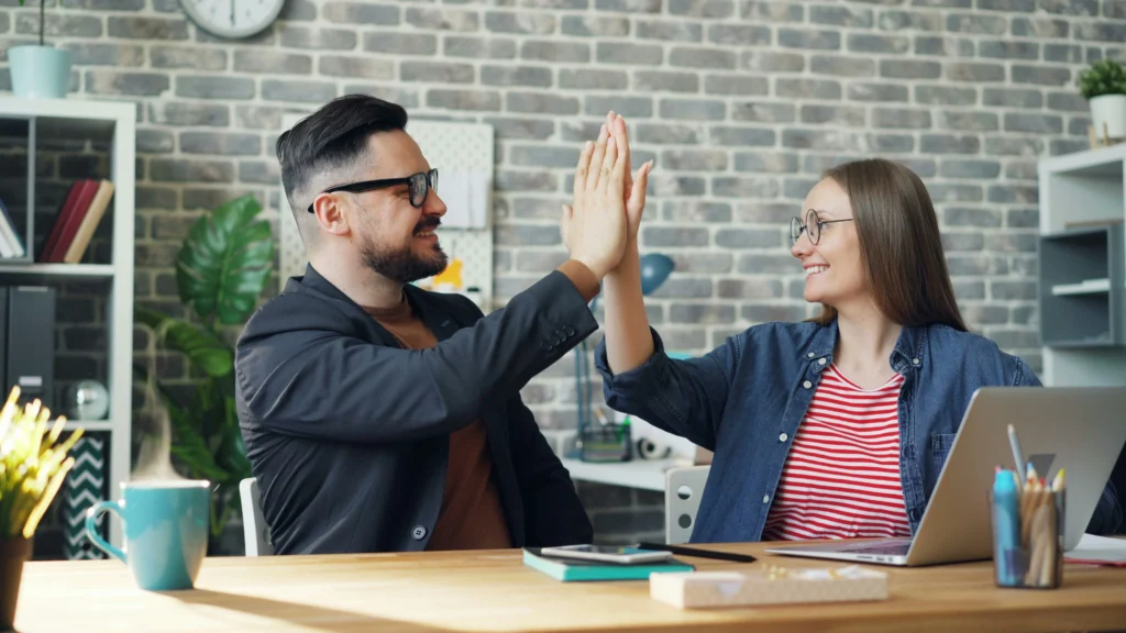 A man and a woman, both wearing glasses, sit across from each other at a wooden desk in a brick-walled office. They are smiling warmly and sharing a high-five over a desk that holds a laptop, stationery, and a steaming blue mug.