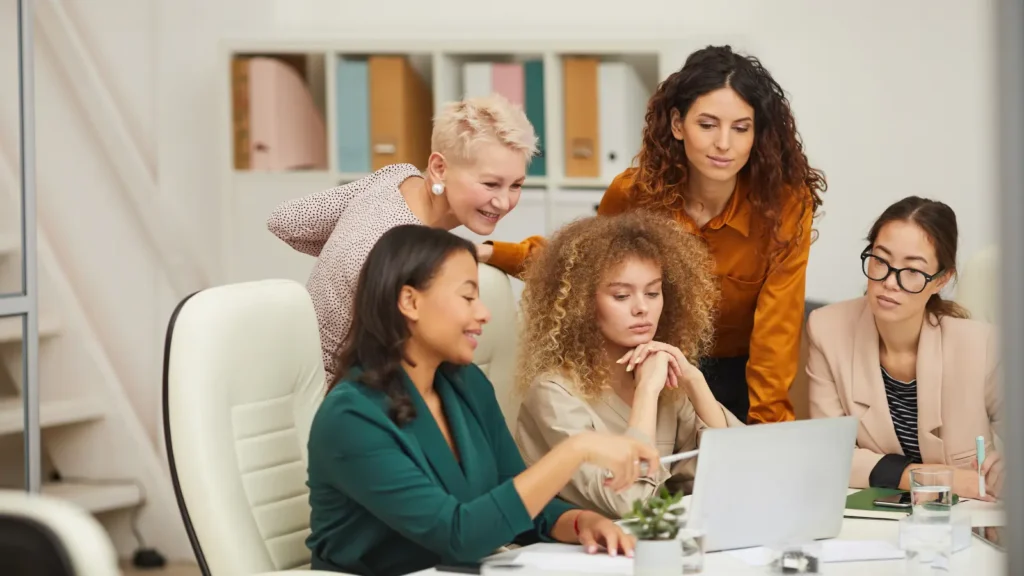 Five diverse women collaborate around a laptop in a bright office, discussing ideas with notes and coffee; warm, focused, friendly teamwork.