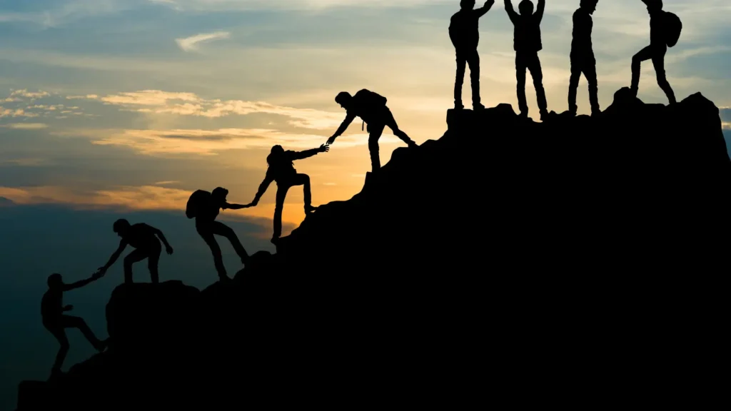 Silhouetted climbers form a human chain up a rocky ridge at sunset, helping one another toward the summit beneath a glowing sky.