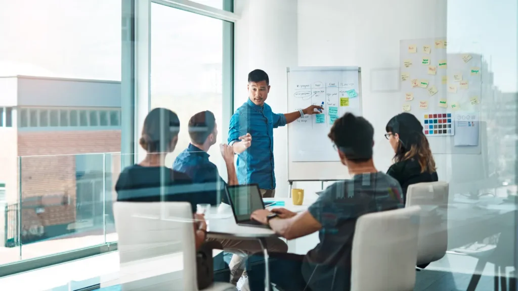 Diverse team collaborates in a bright glass office as a presenter points to a whiteboard while colleagues listen around a table.