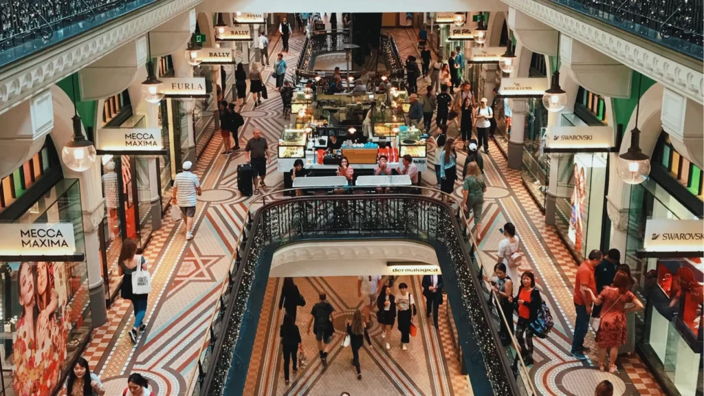 Shoppers stroll through a multi-level historic indoor shopping arcade featuring ornate architectural details, tiled floors, and boutiques.