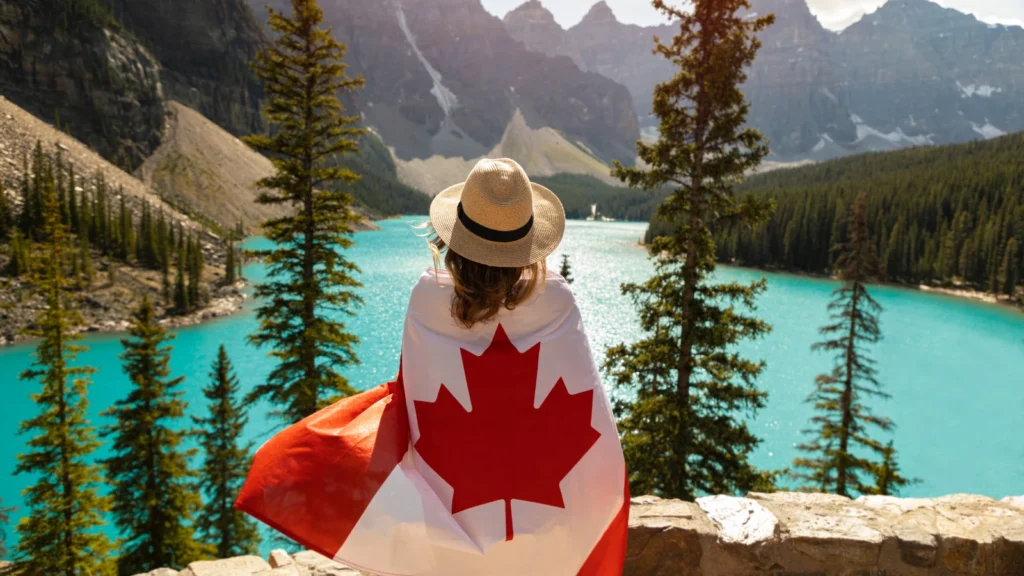 Person wearing a sunhat draped in a Canadian flag, overlooking Moraine Lake and surrounding mountains.