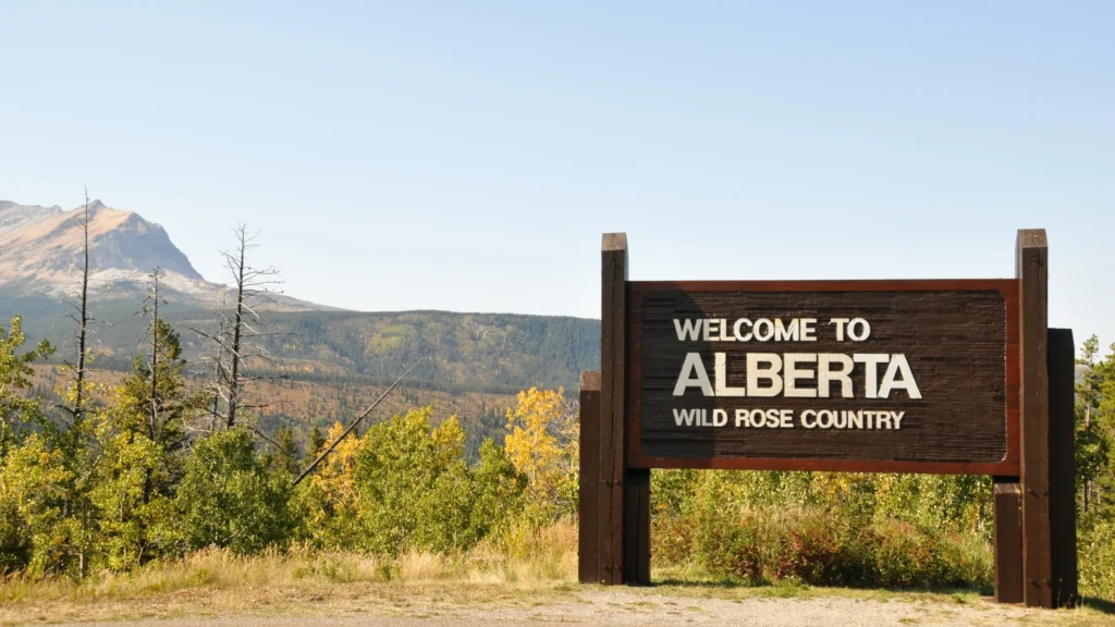Wooden sign reading "Welcome to ALBERTA WILD ROSE COUNTRY" with mountains and forest in the background.
