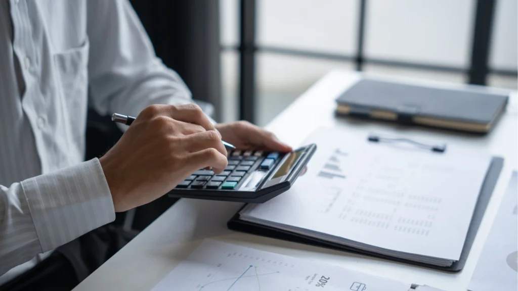 A professional in a white shirt calculates figures on a desktop calculator alongside reports and financial charts.
