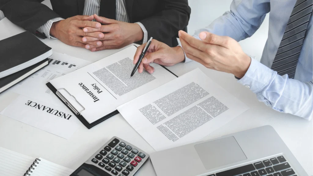Business professionals review insurance paperwork at a desk, pointing with a pen beside a calculator and laptop in a focused, serious meeting.