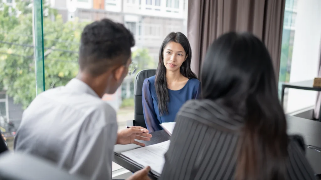 A woman in a blue blouse listens during a professional meeting with two colleagues in an office, conveying a calm, focused tone.