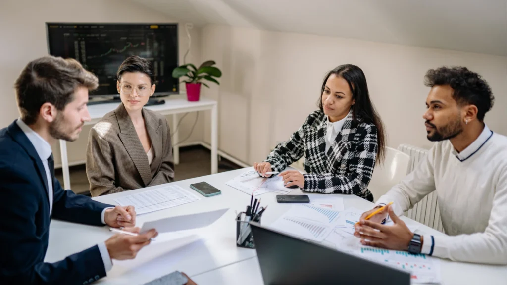 Four coworkers discuss charts and documents around a conference table in a bright office, sharing ideas in a focused, collaborative meeting.