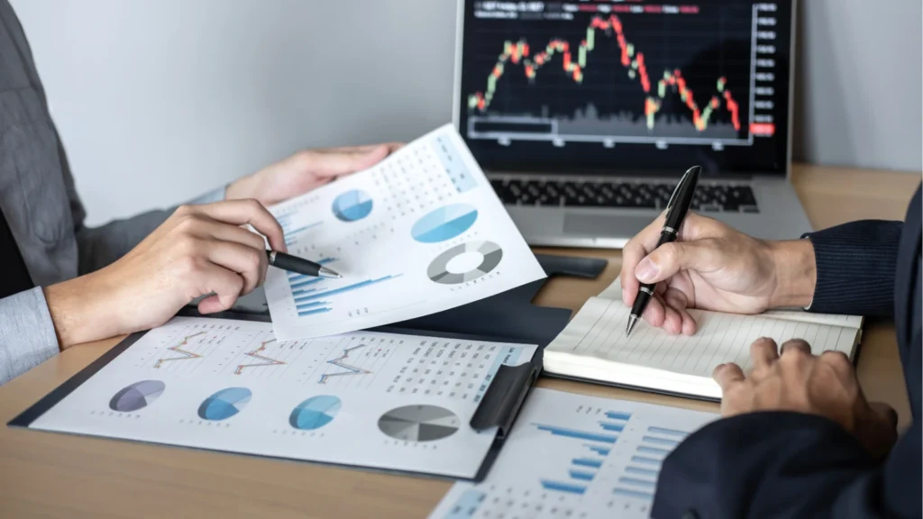 A close-up shot of two professionals collaborating at a desk covered in financial reports and charts. One person points to a pie chart on a printed document, while the other takes notes with a pen next to a laptop displaying live stock market graphs.