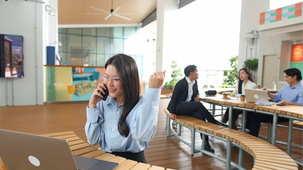 A smiling woman talks on her phone beside an open laptop in a bright modern café, while coworkers chat at a table in the background.