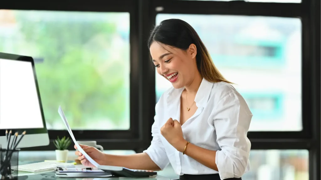 A smiling woman in a bright office reviews paperwork beside a desktop computer, raising a fist in celebration of a work success.
