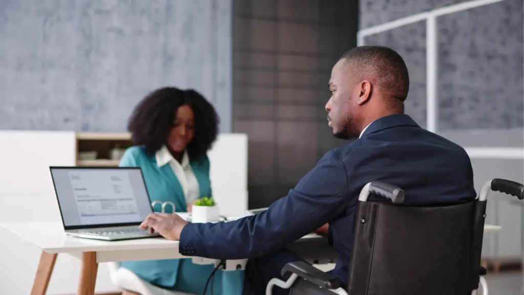 A man in a wheelchair meets with a woman at a laptop in a modern office, suggesting an accessible workplace conversation or consultation.