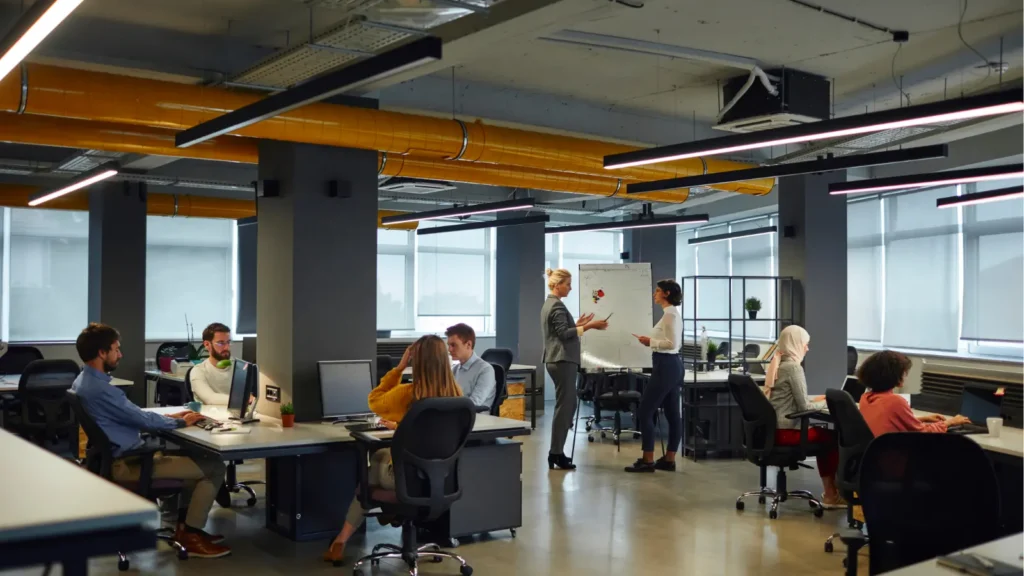 In a bright open-plan office, coworkers work at desks while two colleagues discuss a project beside a whiteboard, creating a calm, collaborative mood.