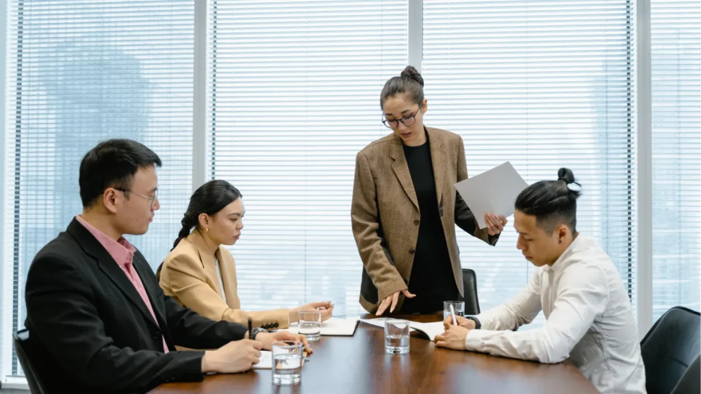 A woman in a brown blazer leads a meeting with three colleagues around a conference table in a modern office.