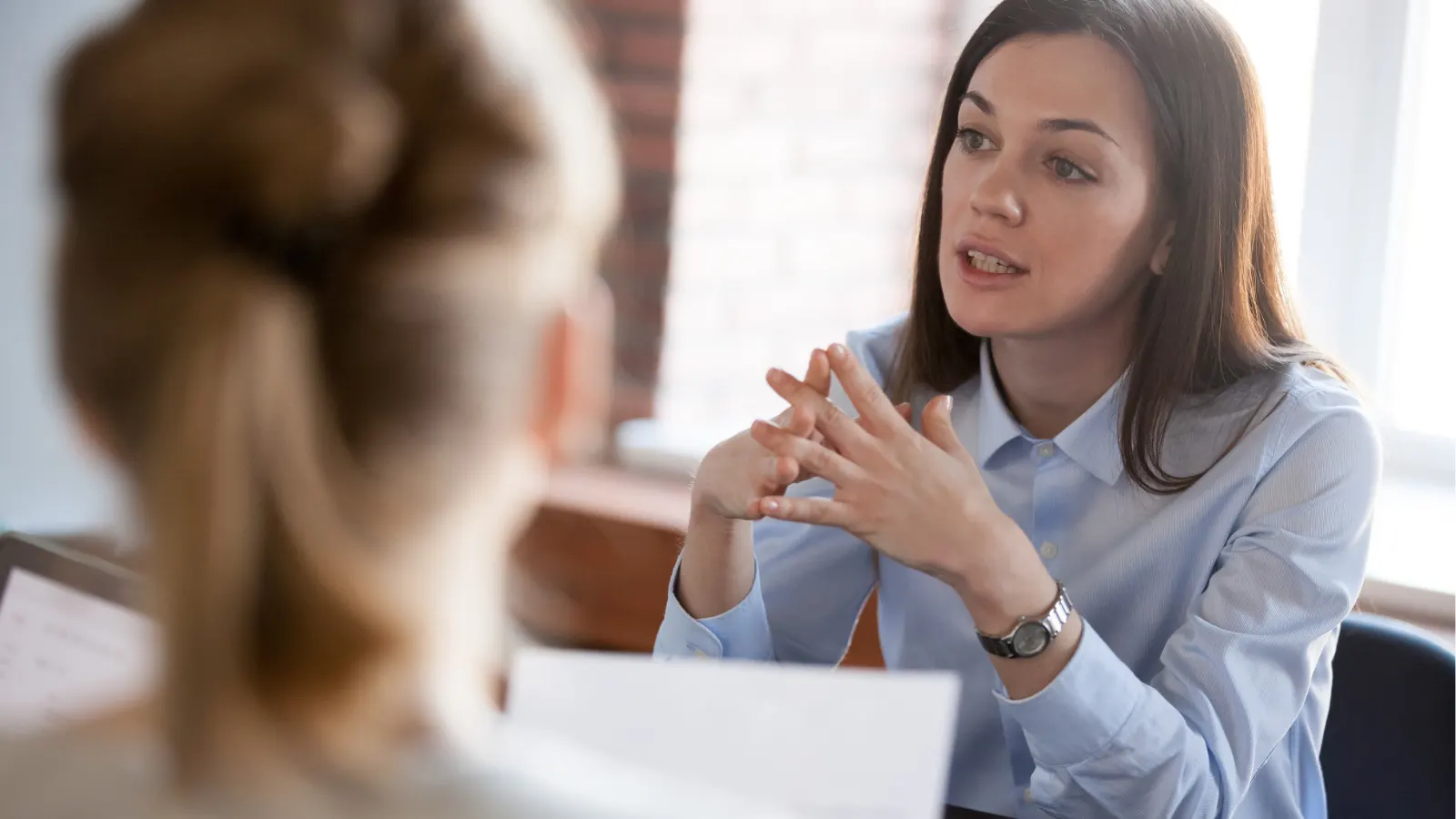A professional woman in a light blue shirt talks passionately during a meeting, gesturing with clasped hands.