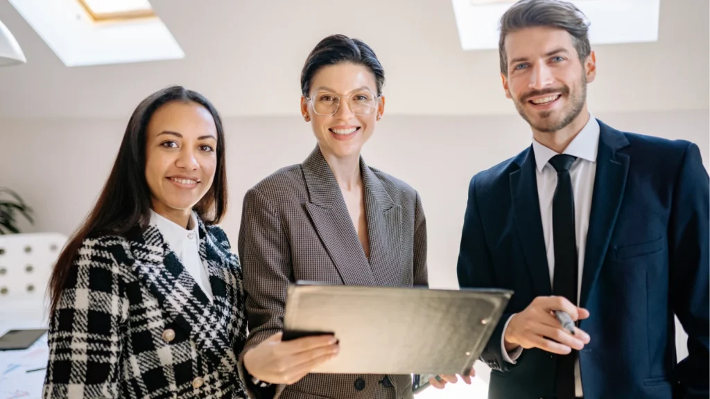 Three smiling professionals standing together in an office while holding a clipboard and pen.