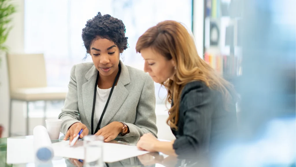 Two female colleagues sitting together at a desk reviewing documents and pointing at paperwork during a business meeting.