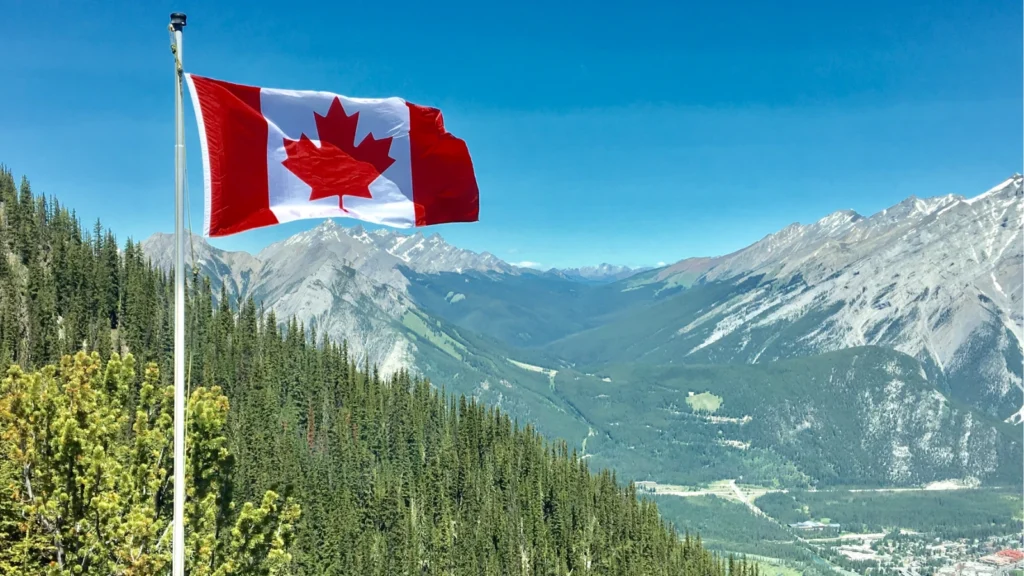 The Canadian flag waving on a flagpole overlooking a lush green forest, valley, and majestic mountain range under a blue sky.