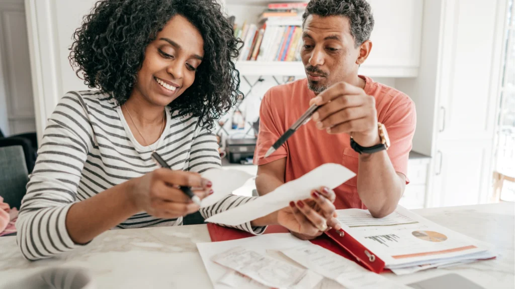 A man and a woman sitting at a table together reviewing personal finance documents and receipts spread over a red binder. Both hold pens and examine paper statements closely.