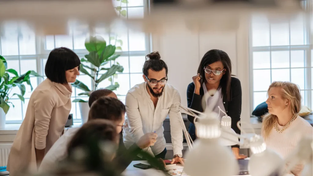 A diverse team gathers around an office table to discuss documents in a bright, plant-filled room, creating a focused, welcoming mood.