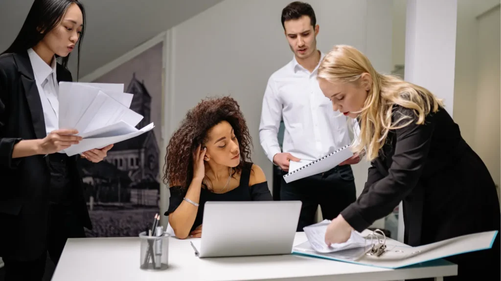 Four coworkers review documents around a laptop in a modern office; a blonde woman points to paperwork as colleagues watch, conveying focused teamwork.
