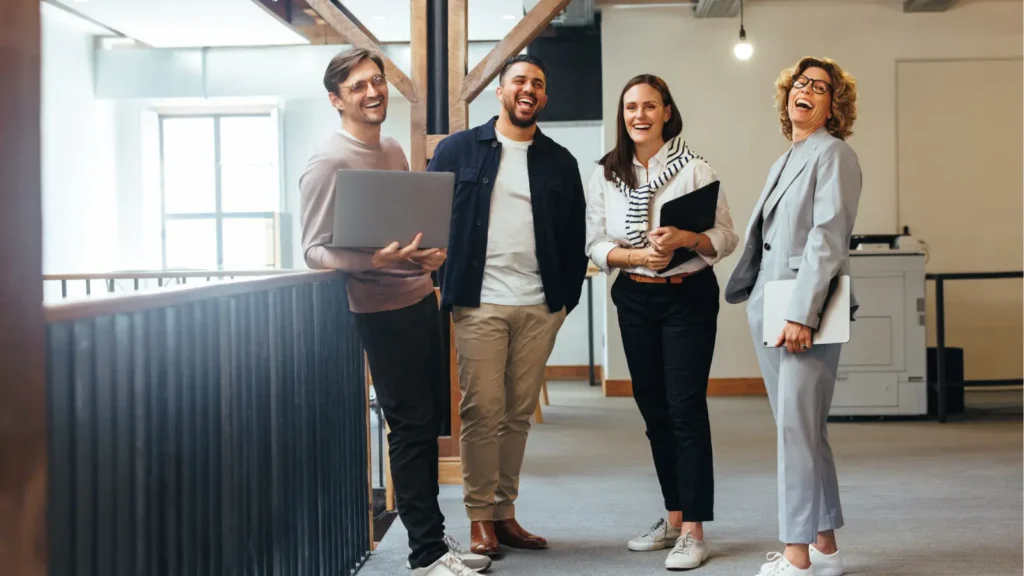 Four diverse coworkers smile in a bright, modern office; one holds a laptop as the team stands together, conveying a warm, upbeat workplace atmosphere.