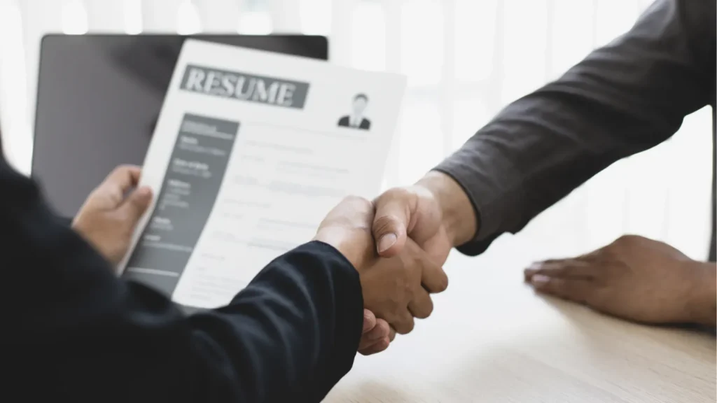 Two professionals shake hands across a desk while one holds a printed resume document