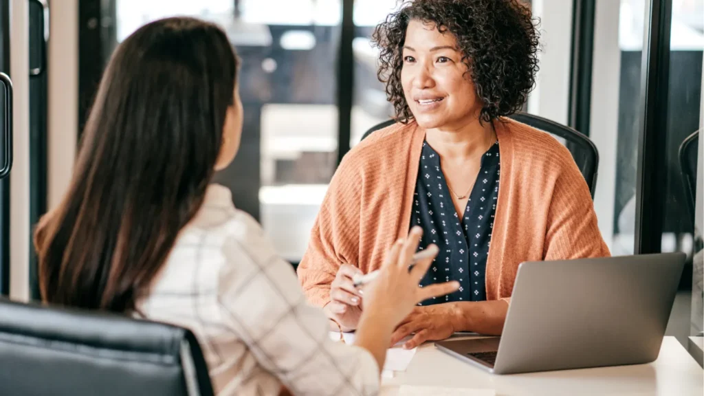 A curly-haired interviewer listens to a job applicant during an interview at an office desk with a laptop.