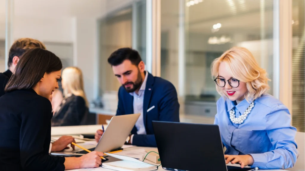 A group of office professionals work collaboratively around a large white table with laptops, featuring a blonde woman with round glasses and red lipstick typing on a dark laptop in the foreground.