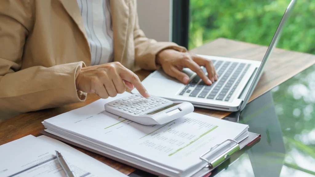Close-up of a person at a wooden desk using a white calculator and laptop over financial papers.