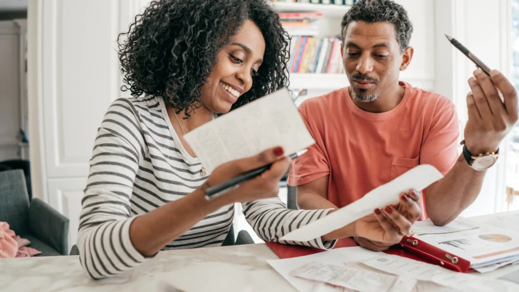A smiling couple sitting together at a table, reviewing paper bills and financial documents.